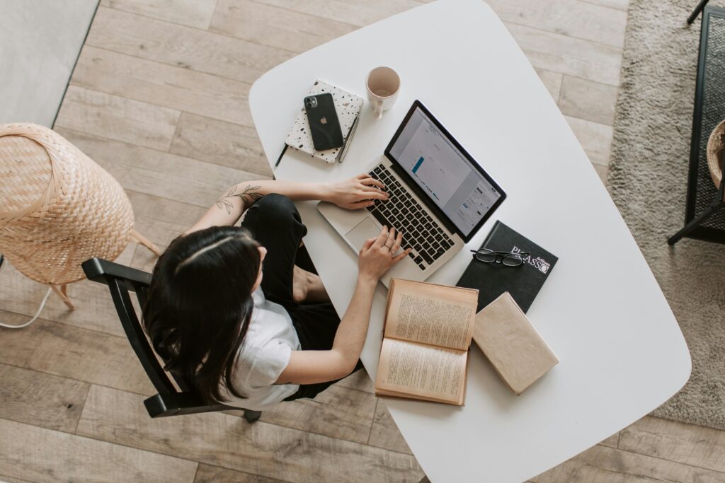 Overhead view of a woman using a laptop at a home desk, surrounded by books, a phone, and a cup.